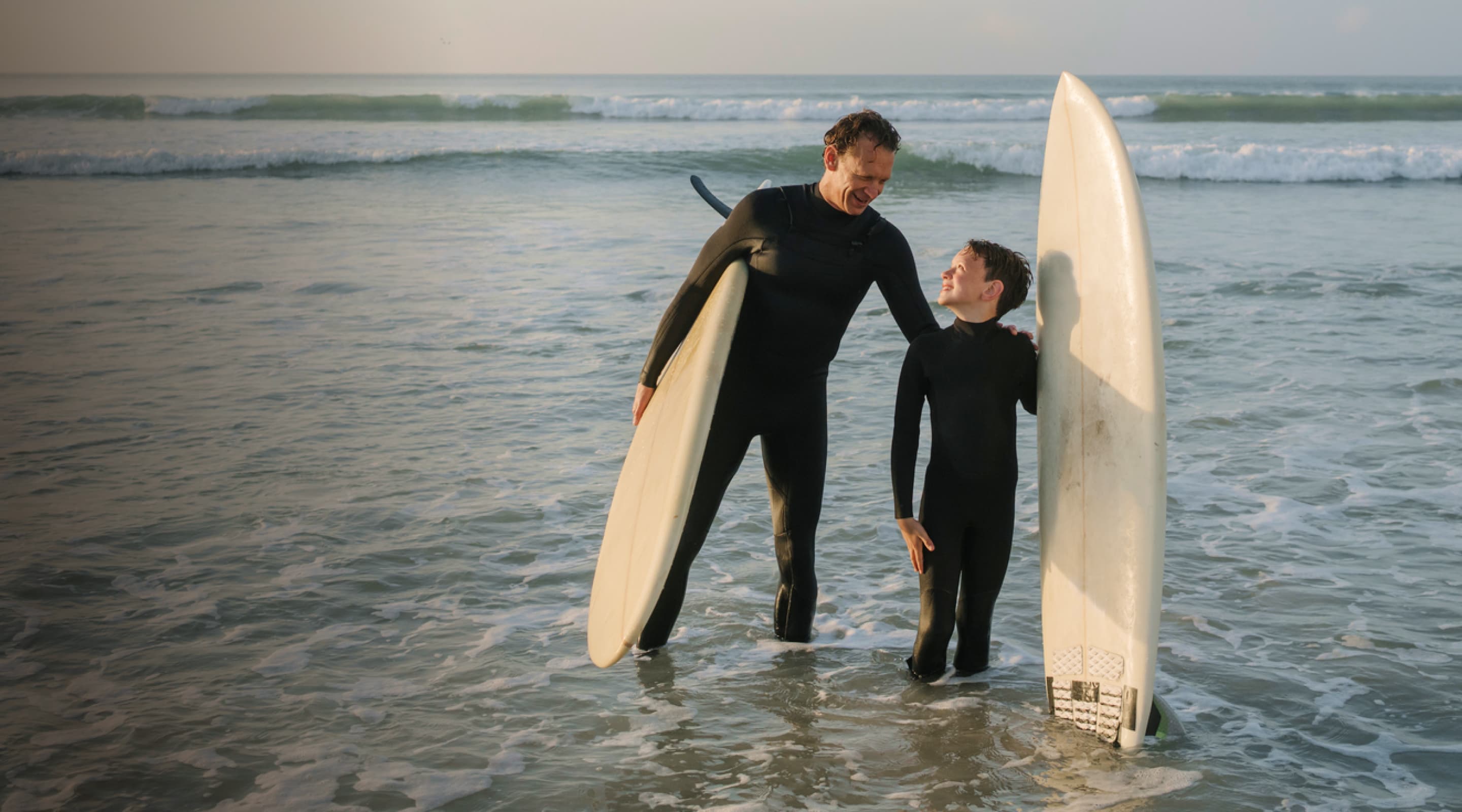 A man and a child in wetsuits stand in the ocean, holding surfboards and smiling at each other with waves in the background.
