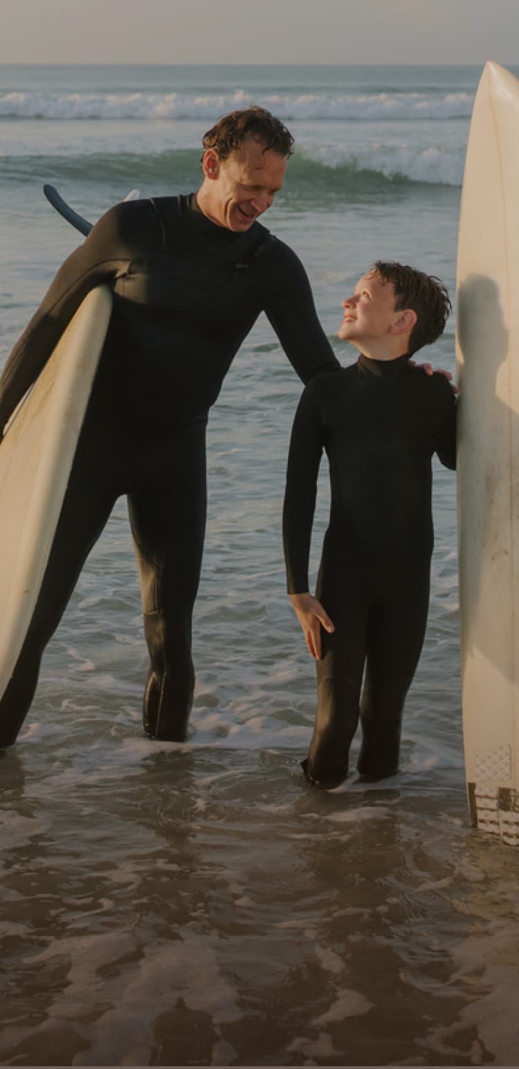 Two people in wetsuits stand in shallow ocean water, holding surfboards, smiling at each other against a backdrop of waves and a clear sky.